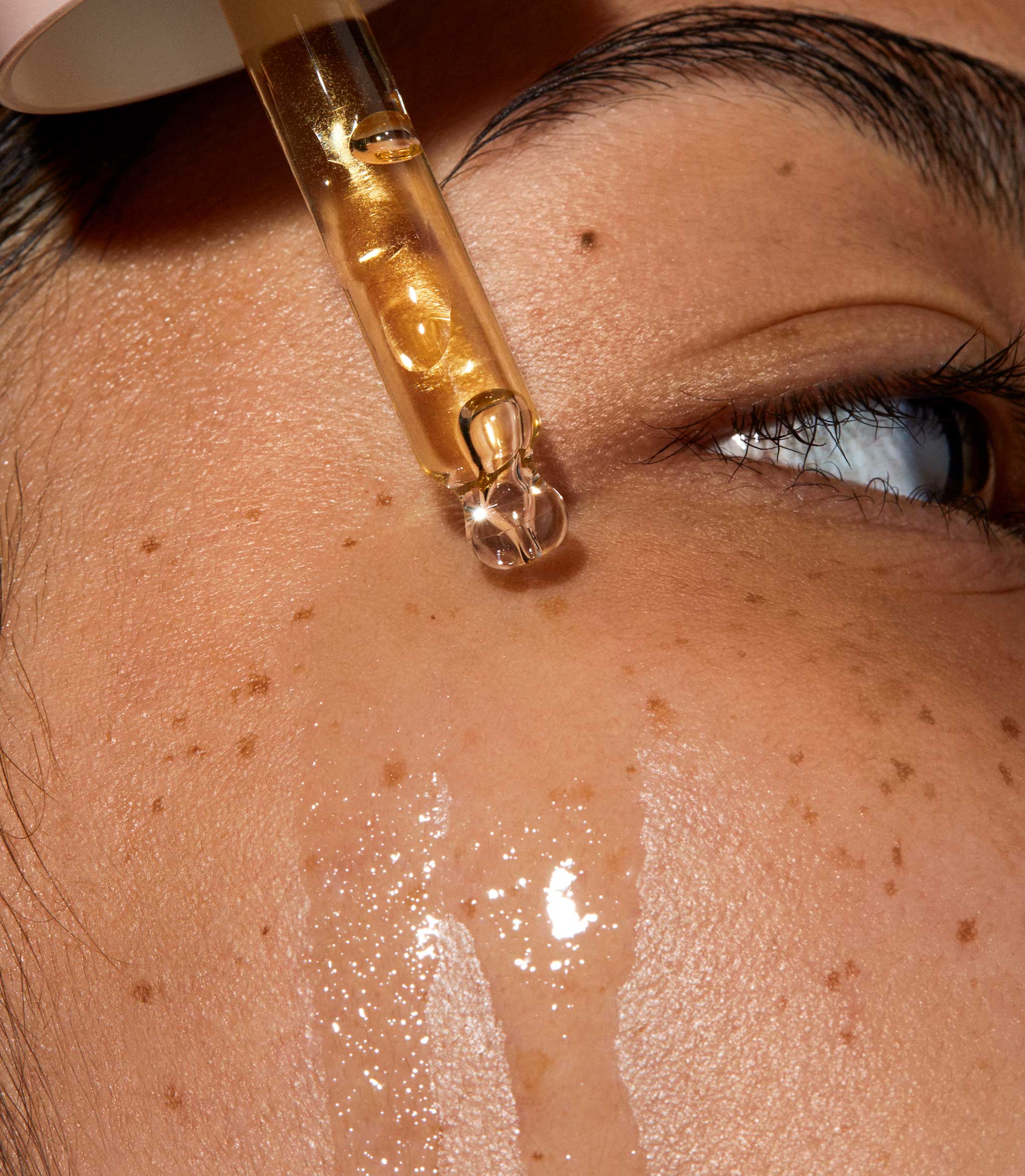 A super close up of model's face Queen oil is applied to her skin with a pipette. The skin is glowy and hydrated.