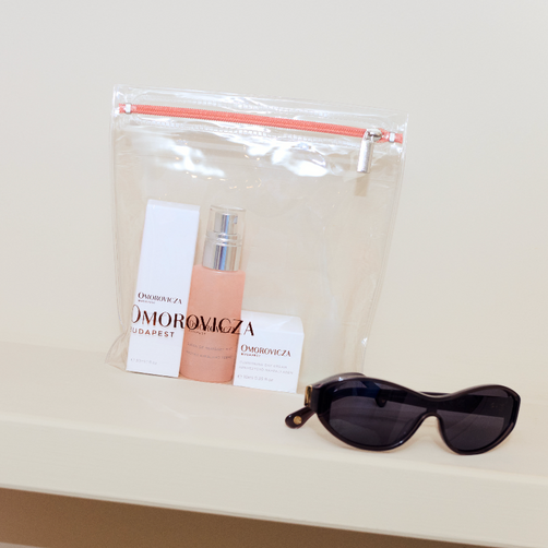 A clear zippered pouch containing a small beige bottle and two white cards with the brand name Omoromicza, displayed on a cream shelf next to black sunglasses.