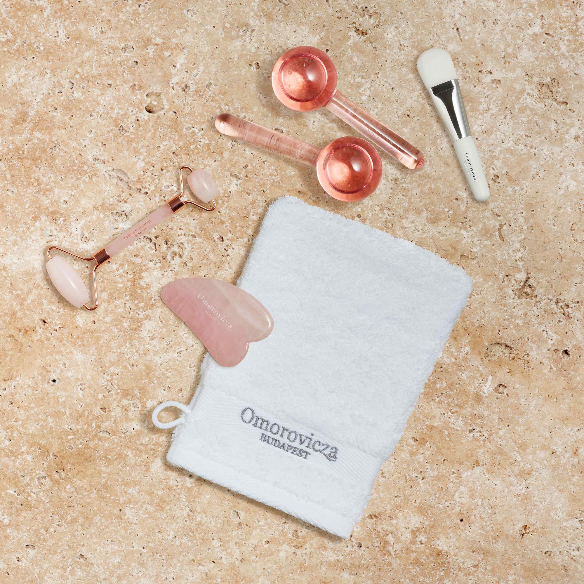 White cleaning Mitt with Omorovicza logo on a pink stone next to white Mask Brush, Cooling Dermaglobes, Rose Quartz Roller and Sculpting Rose Gua Sha.
