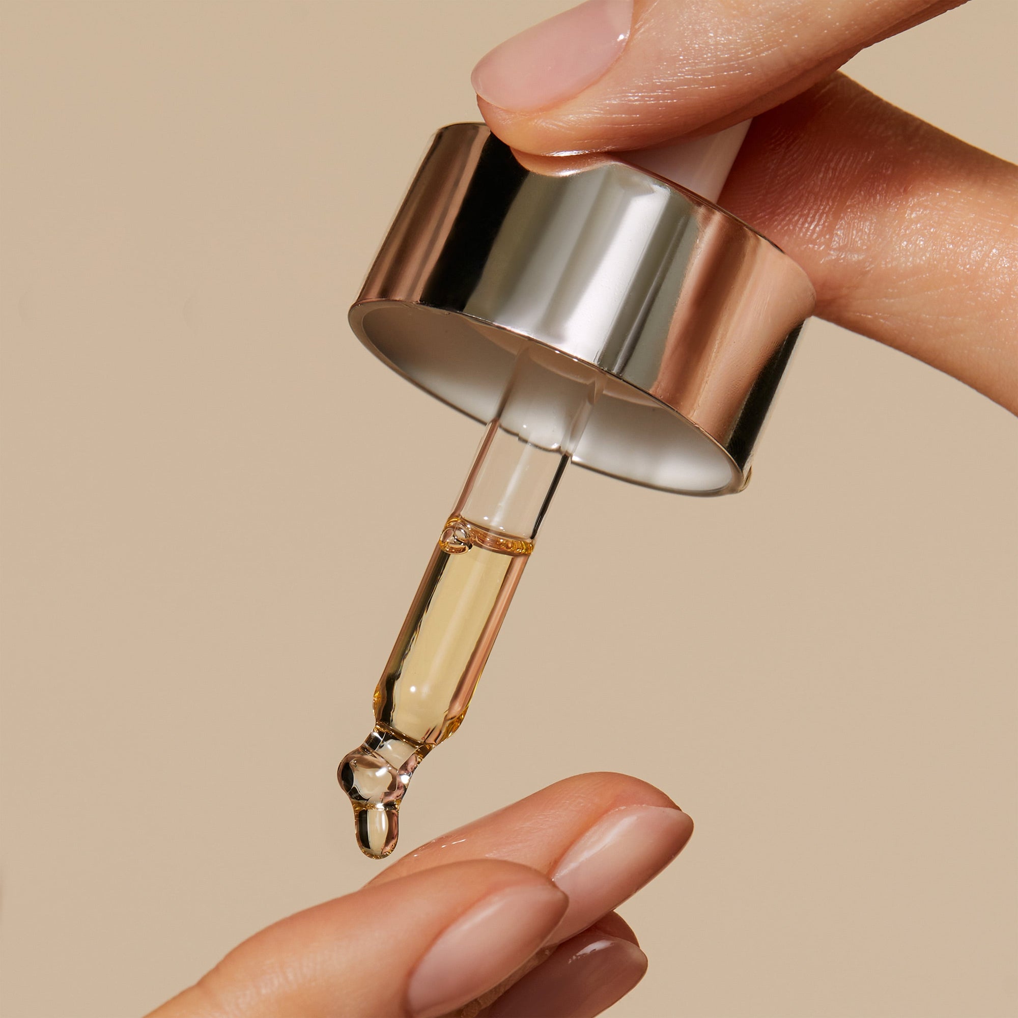 Hand is holding a pipette with Miracle Facial Oil. the drops of oil are falling on models fingers. The texture of the oil is rich and has a tint of gold colour.